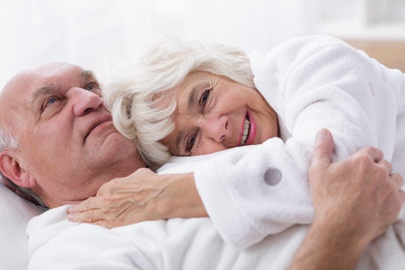 Older couple lying on a bed, smiling and embracing.